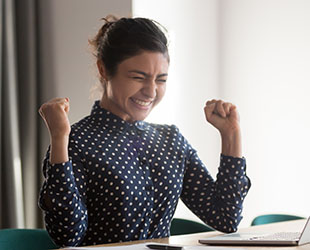 Excited young woman fist pumps while looking at computer screen. Happy job seeker receives offer from employer
